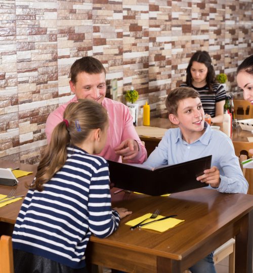 Adult hospitable waitress helping visitors with menu taking order in cafe