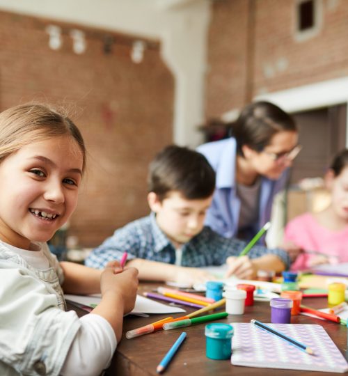 Portrait of cute little girl looking at camera while enjoying art class with group of children, copy space